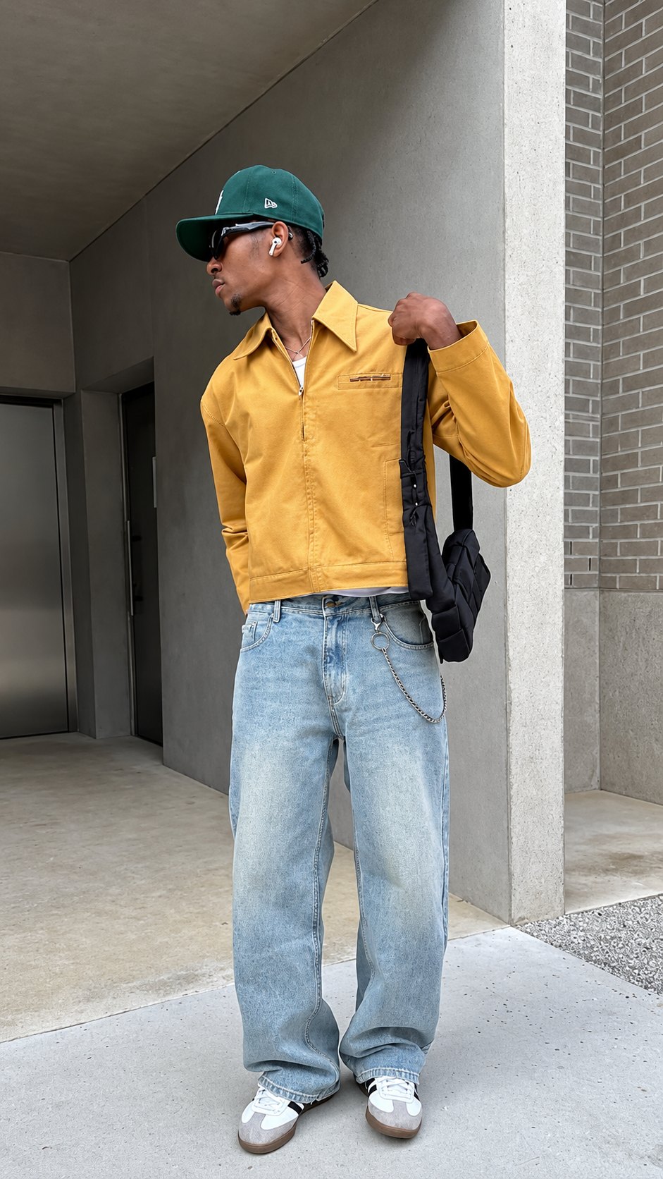 A man in a green cap, yellow jacket, light baggy jeans and white sneakers